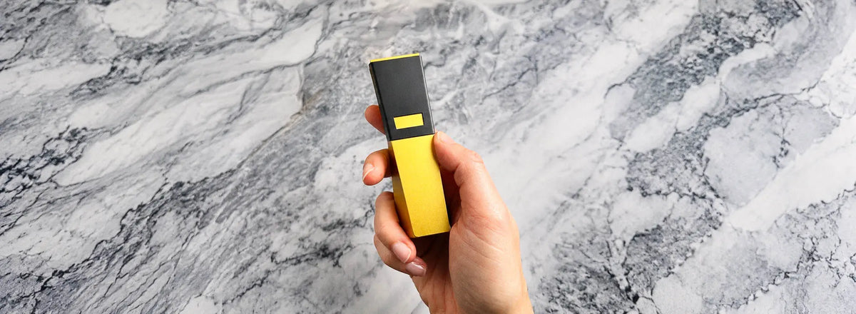 Hand holding gold Tempest Radiance smoking pipe with black magnetic Tempest Shield bowl cover against white marble background