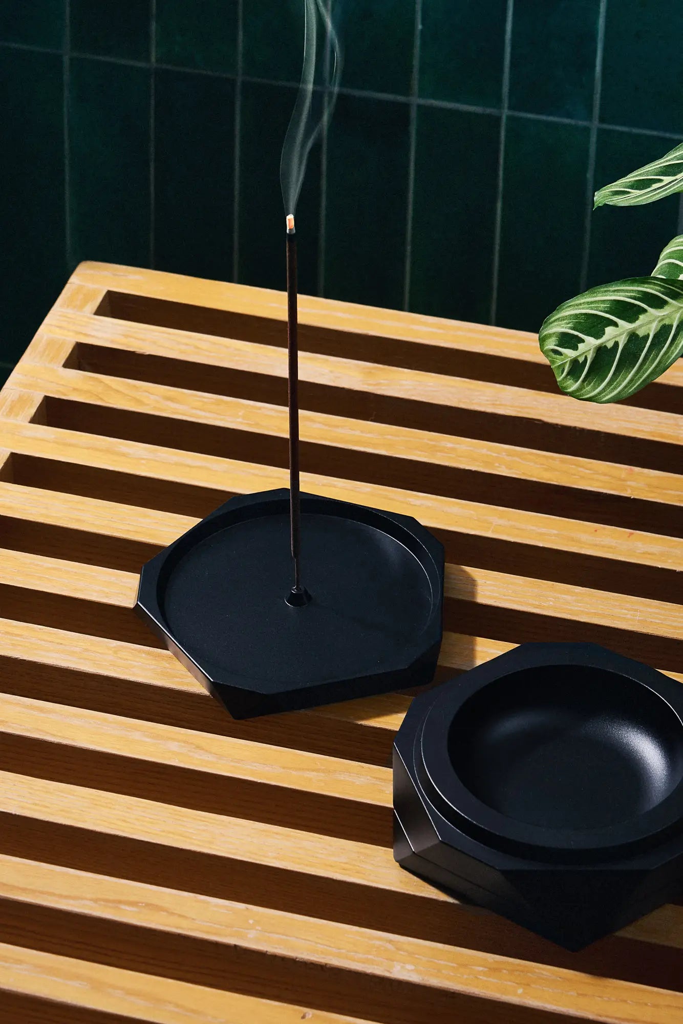 Incense stick burning in a black dish on a wooden surface with a dark green tiled wall in the background.